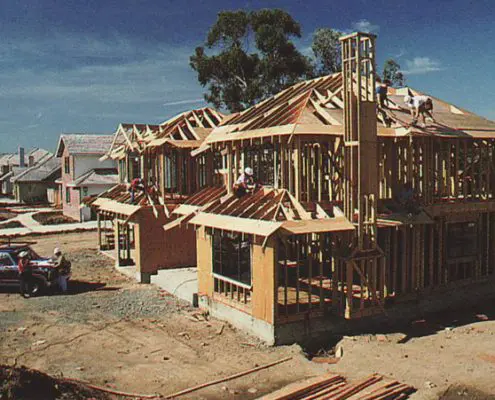 Two-story wooden house under construction with L.E. Baca Construction workers on the roof