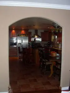View through arched doorway into custom kitchen with wood cabinetry, stainless steel appliances, and tiled floor – L.E. Baca Construction project