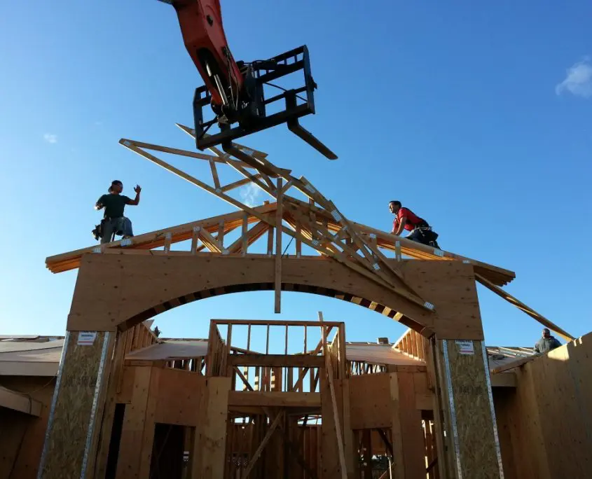 New home construction with an arched entryway below and workers on the roof placing large timber trusses