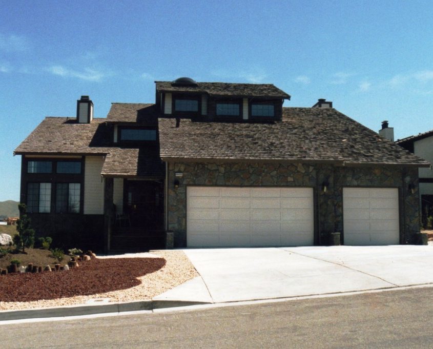 A large, modern house with a light beige or tan exterior, dark brown trim, and a prominent stone facade around the two-car garage.