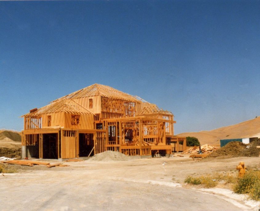 New home construction showing exposed wooden studs and roof trusses, surrounded by excavated dirt and construction materials