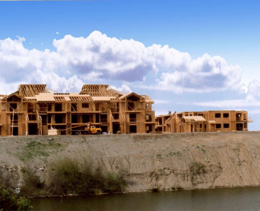 Multiple townhouses under construction, framed with wood, sit atop a dirt embankment overlooking a body of water under a partly cloudy sky